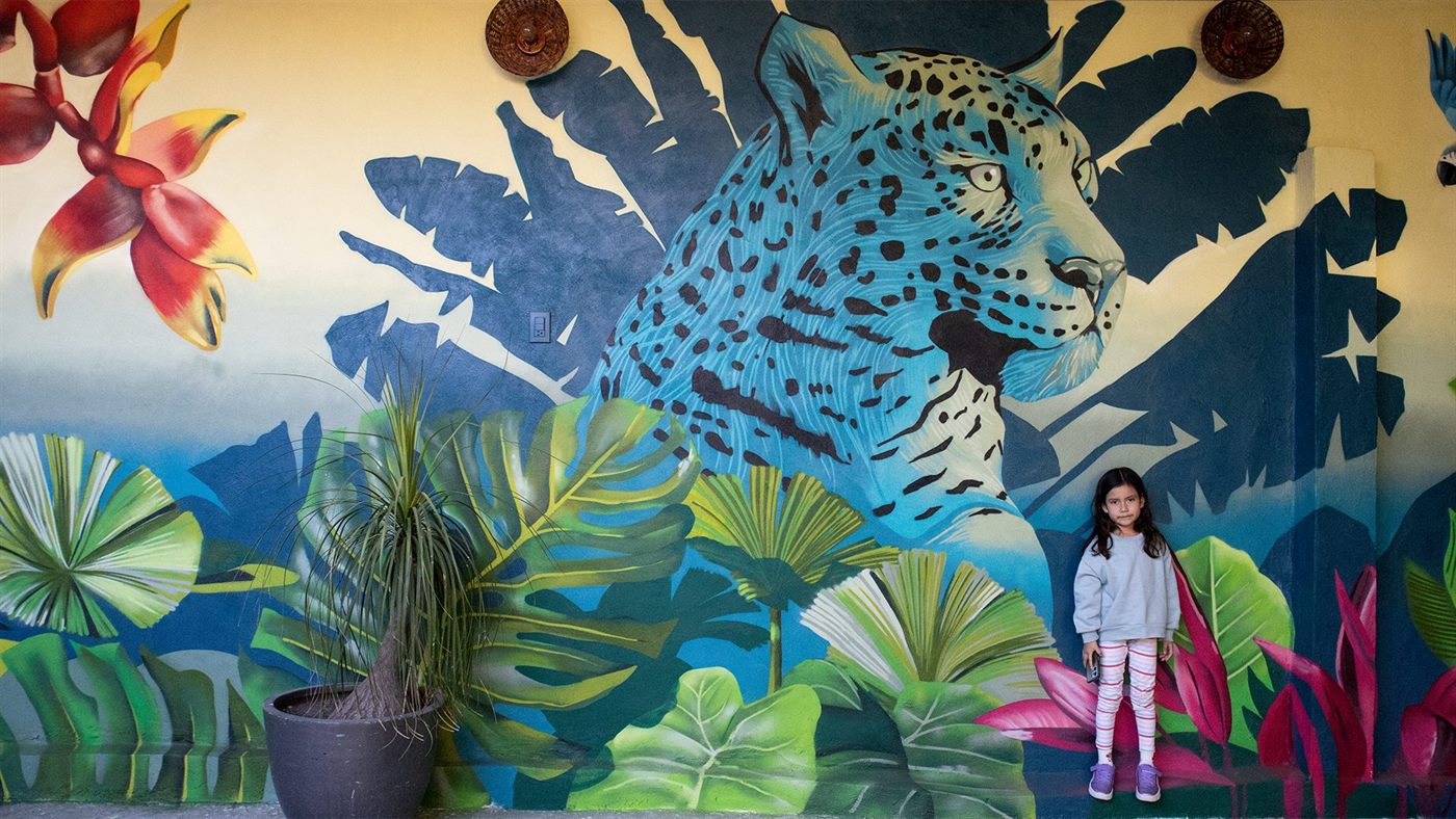 Una niña de pie junto al jaguar del mural, que muestra la escala real de la pieza en la terraza